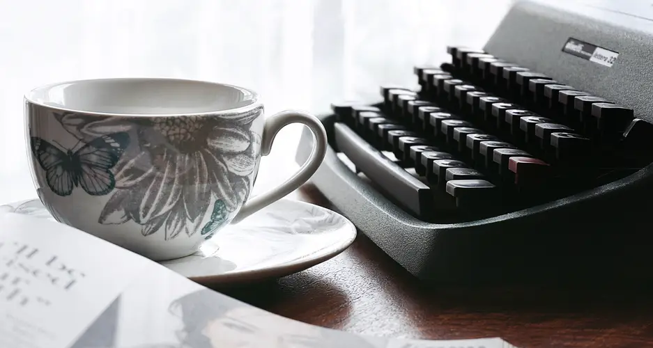 Weiße Tasse mit Schmetterlings- und Blumenmuster auf Untertasse neben alter Schreibmaschine und offenem Buch.